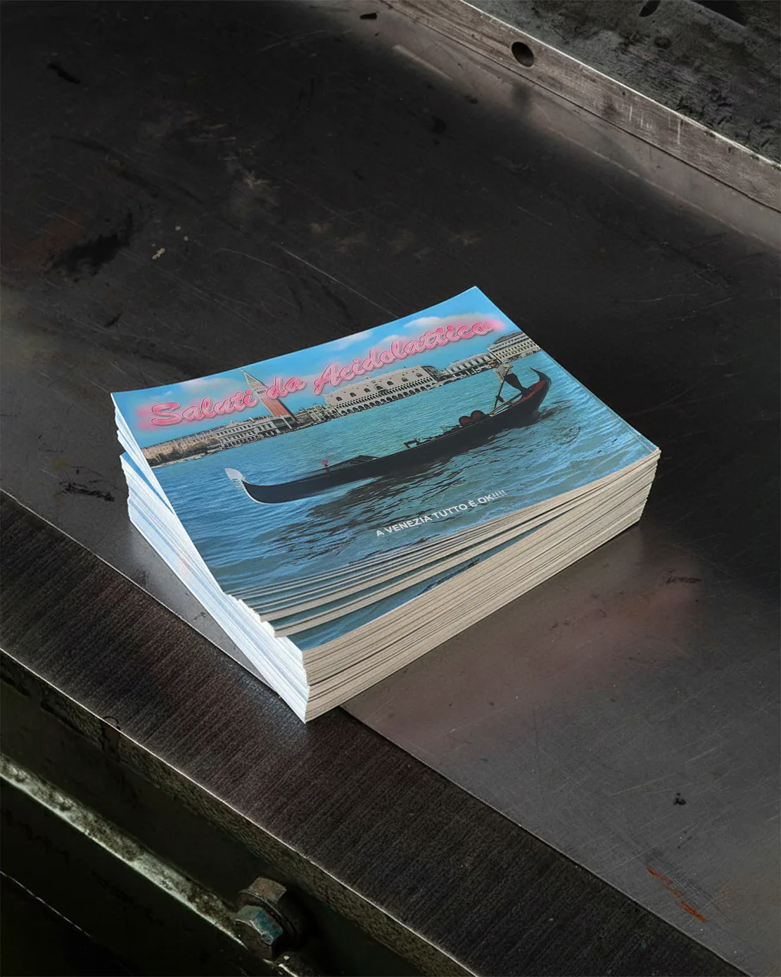 Pila di cartoline “Saluti da Acidolattico” con immagine di gondola a Venezia, poggiata su un piano di metallo scuro.