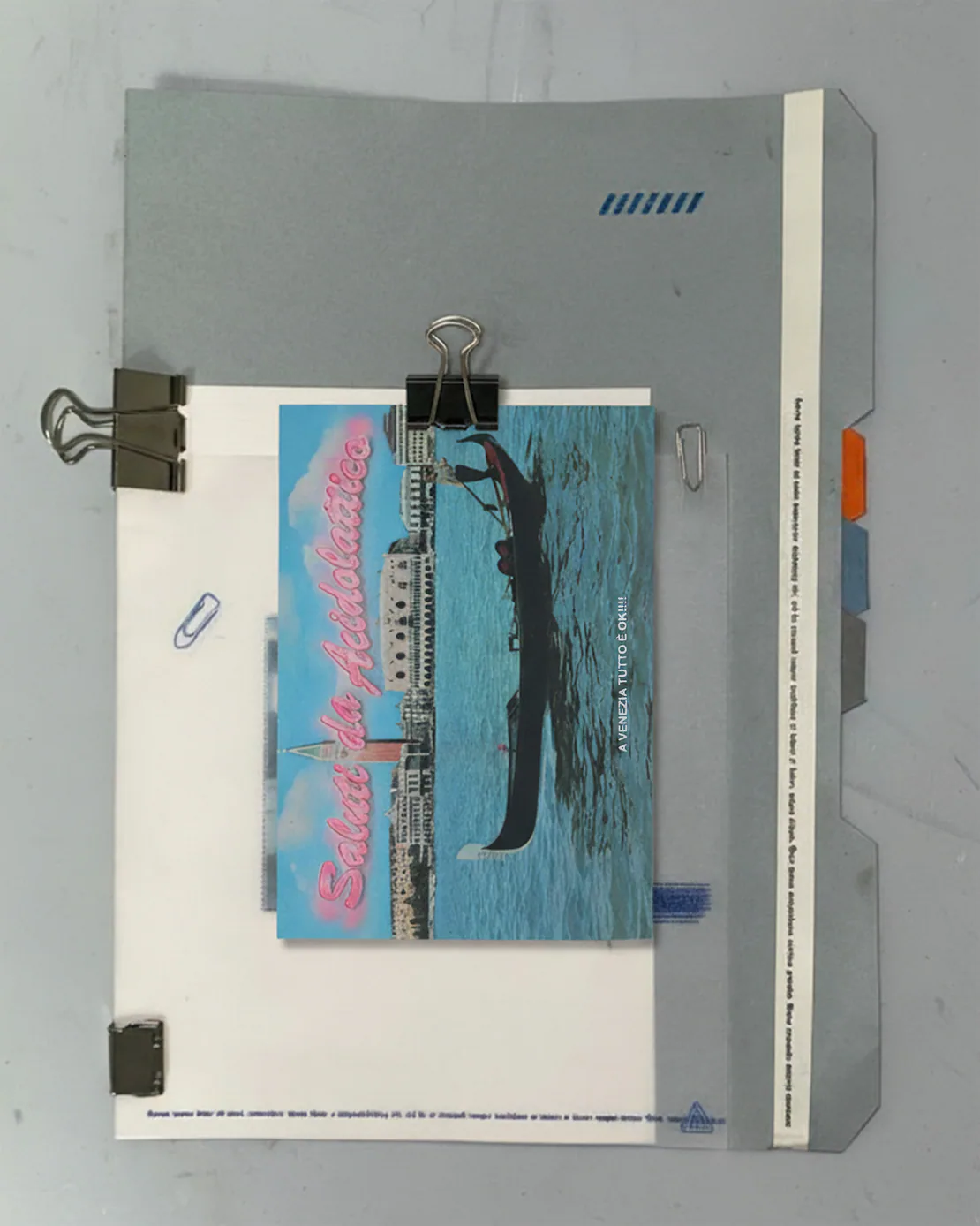 Cartolina “Saluti da Acidolattico” fissata con clip metalliche a una cartellina grigia, su fondo chiaro.