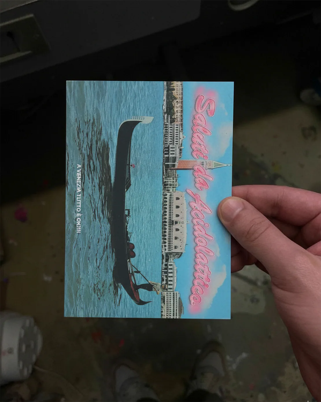 Mano che tiene una cartolina “Saluti da Acidolattico” con gondola a Venezia, su sfondo scuro e pavimento macchiato.
