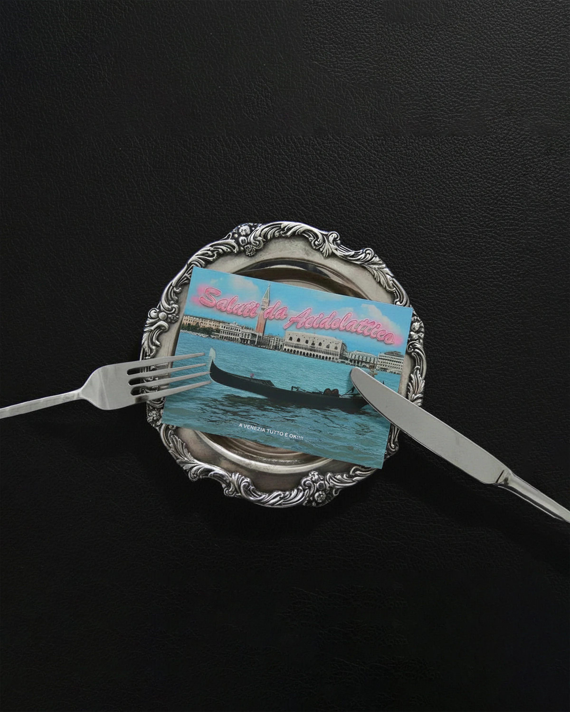Cartolina con la scritta ’Saluti da Acidolattico’ raffigurante Venezia, posata su un piatto d’argento con forchetta e coltello.