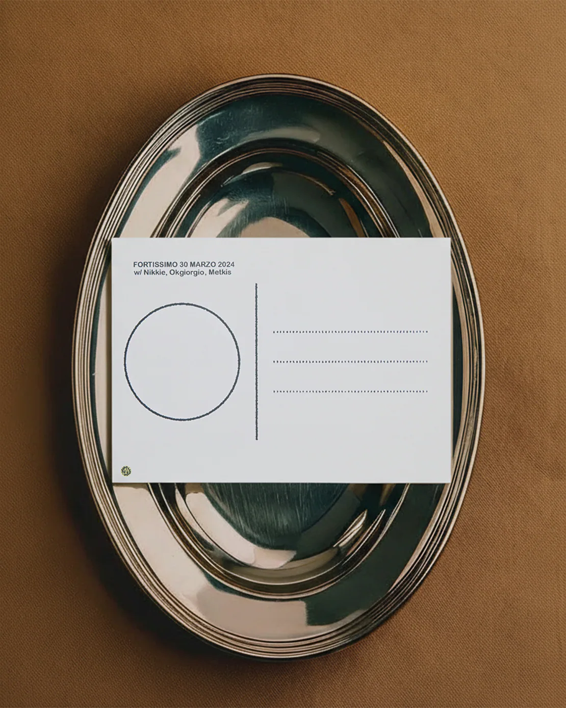 Retro di una cartolina appoggiata su un vassoio d’argento lucido, con testo tipografico nero su sfondo bianco, fotografata su tessuto marrone.