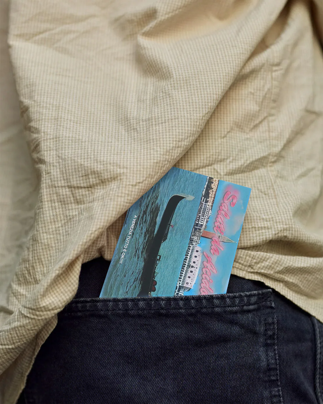 Cartolina “Saluti da Acidolattico” sporgente dalla tasca posteriore di un paio di jeans scuri, con camicia chiara sopra.