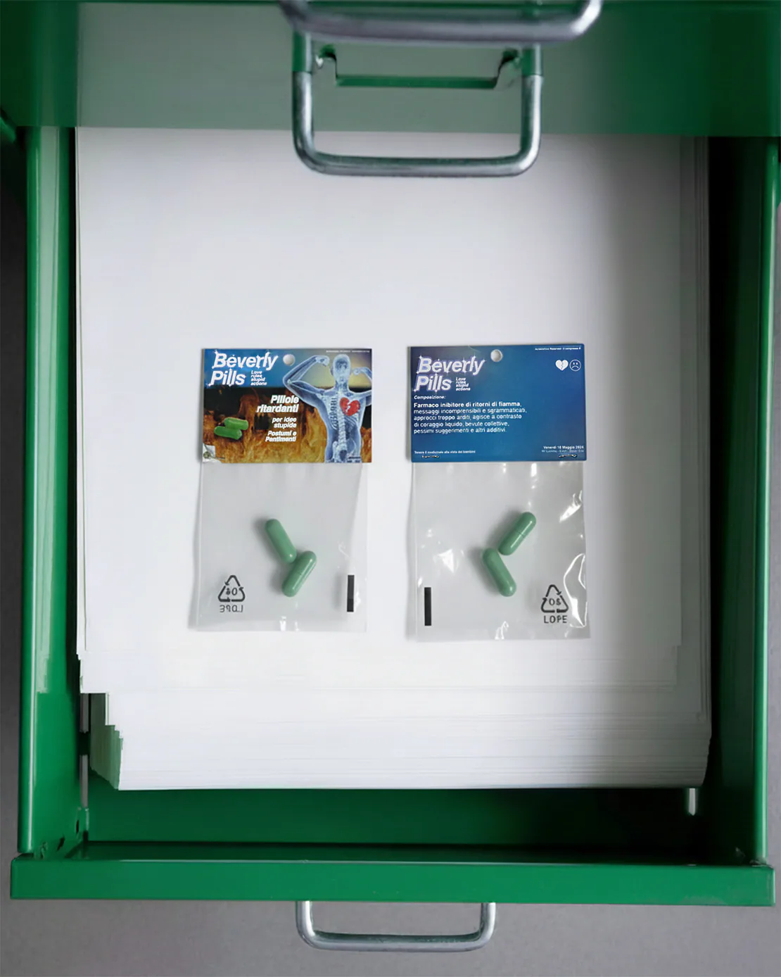 Due bustine di Beverly Pills in un cassetto metallico verde, adagiate sopra una pila di fogli bianchi.
