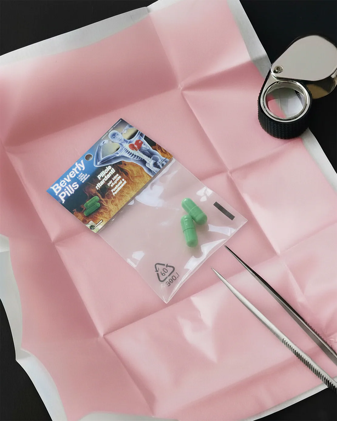 Una confezione di Beverly Pills posata su un foglio rosa piegato, con lente e pinzette da laboratorio accanto.
