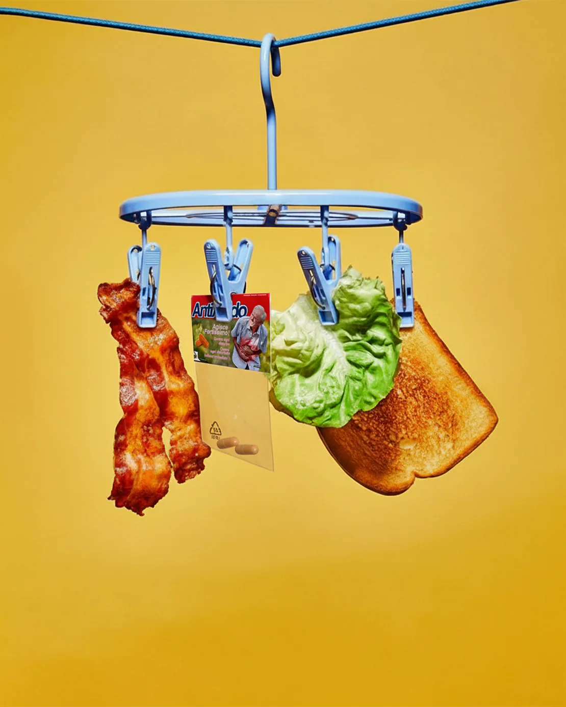 Bustina di Antiacido Pills appesa a uno stendino insieme a pancetta, lattuga e pane tostato, su sfondo giallo brillante.