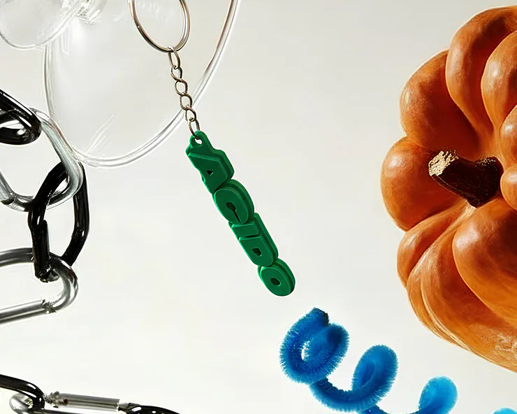 Composizione di oggetti sospesi: moschettoni metallici, bicchiere trasparente, zucca arancione e portachiavi verde con la scritta ACIDO.