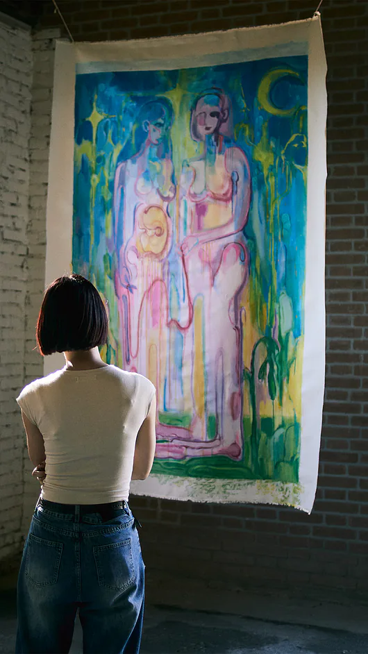 Una donna di schiena osserva un grande dipinto colorato appeso a un muro di mattoni in una mostra di arte.