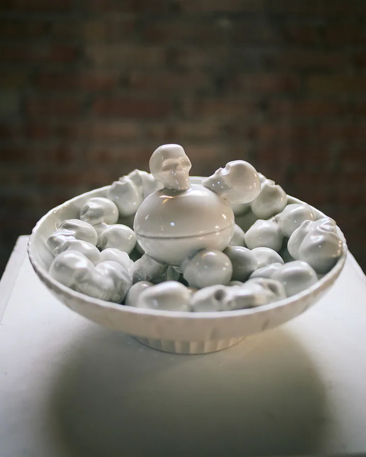Dettaglio di una scultura in ceramica bianca, una ciotola piena di piccole forme rotonde e teschi.