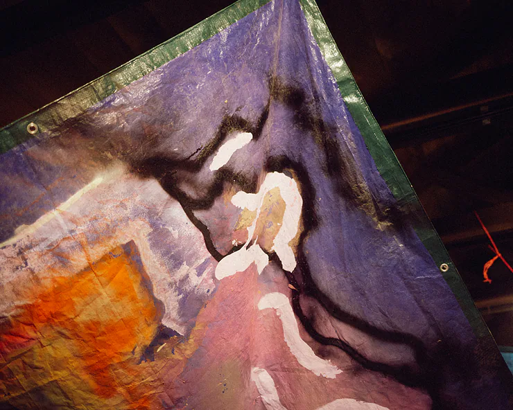 Dettaglio di una opera di arte dipinta su un telone, con colori viola, arancioni e forme astratte in bianco e nero.