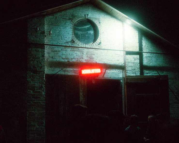Ingresso di un edificio in mattoni di notte, con una insegna rossa al neon con la scritta Acidolattico che illumina l'entrata.