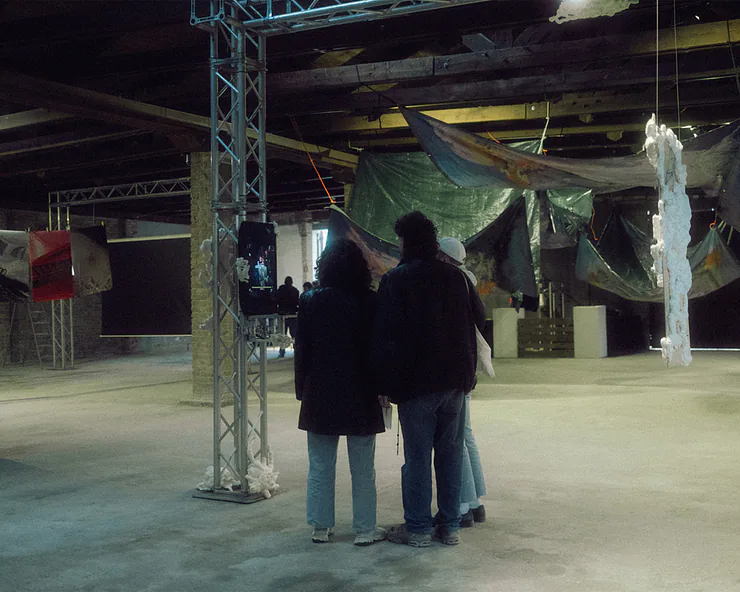 Un gruppo di visitatori osserva le installazioni artistiche in un ampio spazio espositivo industriale.