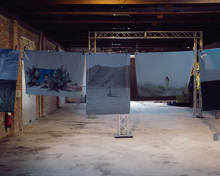 Allestimento di una mostra fotografica con grandi stampe appese a una struttura metallica in uno spazio industriale.