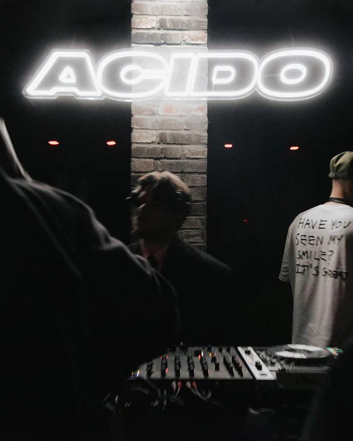 Insegna luminosa con la scritta Acido sopra la console di un DJ durante una serata in un locale buio.
