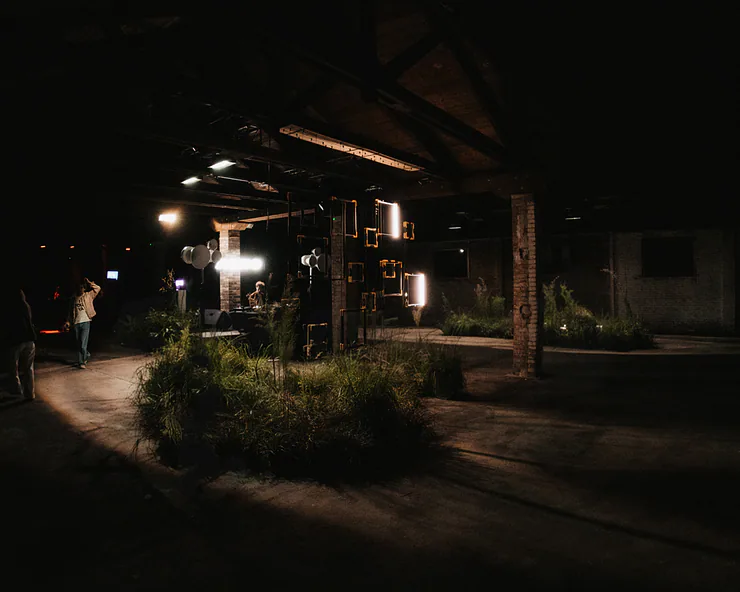Allestimento di un evento in un grande spazio industriale, con isole di erba alta, una console da DJ e installazioni luminose.