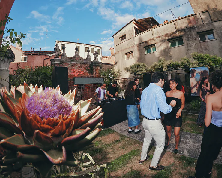 Festa diurna in un giardino a Venezia con persone che ballano vicino alla postazione del DJ. In primo piano un grande fiore di carciofo viola.