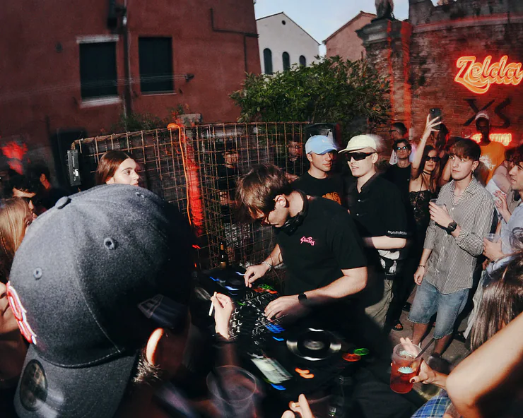 Un DJ suona alla console durante una festa in un cortile a Venezia, circondato da persone. Sullo sfondo si vede una insegna al neon con la scritta Zelda.