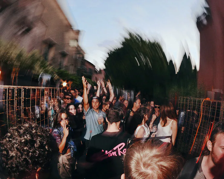 Folla di giovani a una festa in un giardino a Venezia con le mani alzate. La immagine ha un marcato effetto di motion blur.