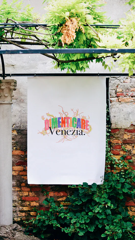 Un poster bianco con la scritta colorata Dimenticare Venezia appeso a un muro di mattoni antico coperto di edera.