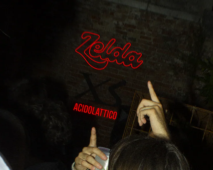 Insegne rosse al neon di Zelda e Acidolattico su un muro di mattoni di notte, con delle mani in primo piano che le indicano.