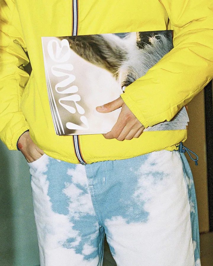 Persona con una giacca gialla e jeans tie-dye che tiene sotto braccio una copia della rivista Lamb durante un evento.