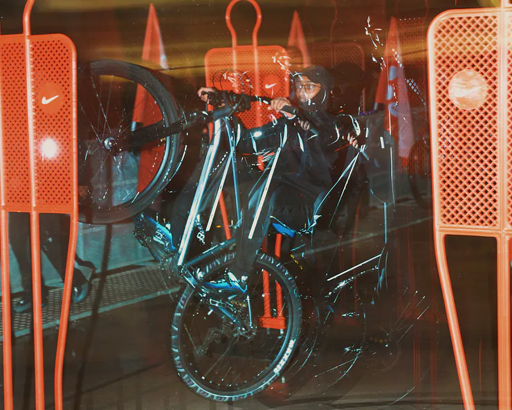 Foto artistica con effetto mosso di un ragazzo che fa una impennata in bici, la cui sagoma è visibile attraverso degli ostacoli da allenamento arancioni con il logo Nike.