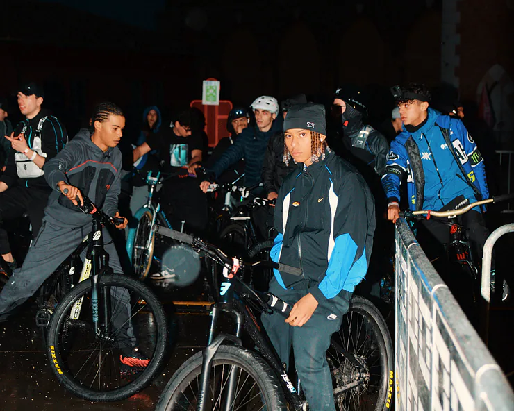 Un gruppo di giovani ciclisti in abbigliamento sportivo e streetwear, inclusi capi Nike, posano con le loro biciclette durante un evento notturno.
