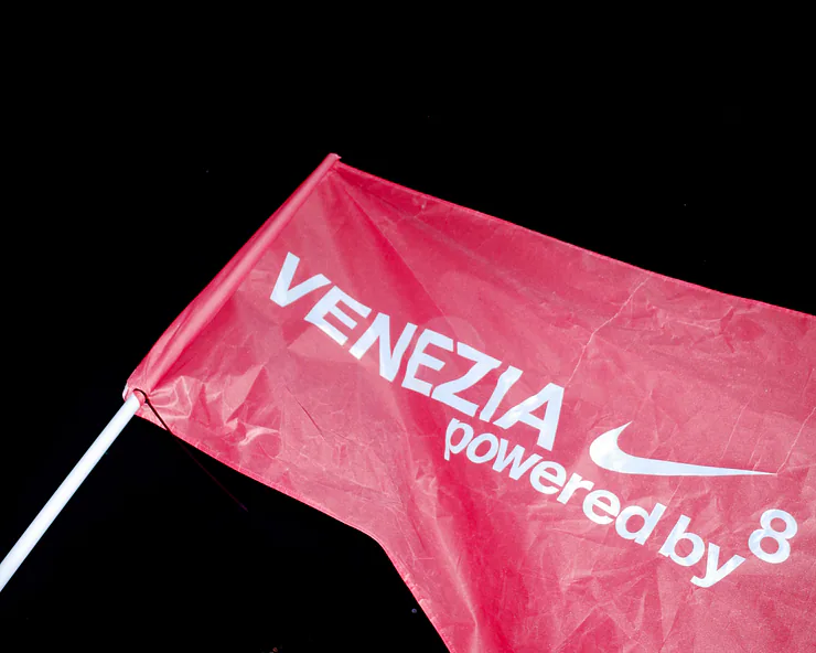 Primo piano di una bandiera rossa del evento VENEZIA 8 powered by Nike con il logo Nike e il testo in bianco su sfondo nero.