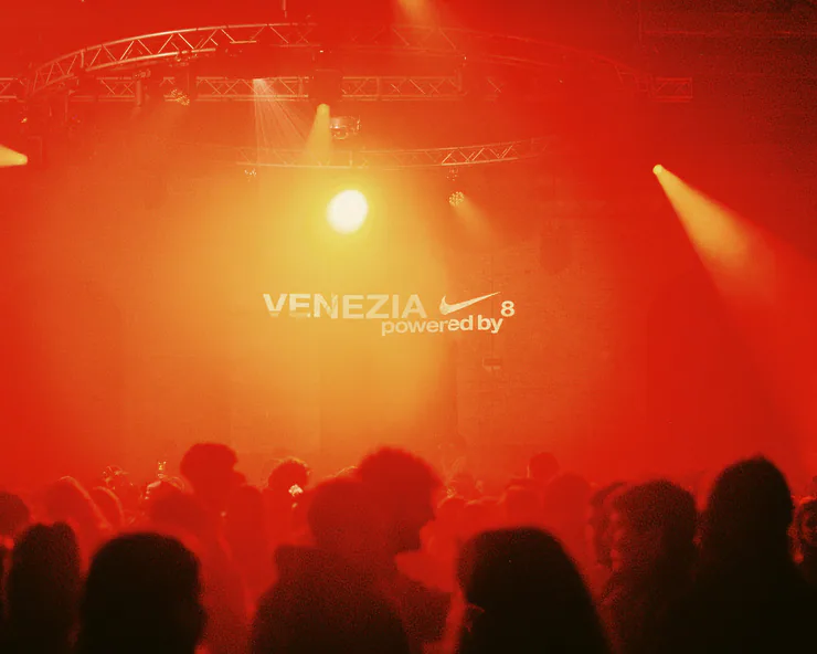 La folla in controluce a un evento serale illuminato di rosso. Sul muro campeggia il logo VENEZIA 8 powered by Nike, indicando la sponsorizzazione del evento.