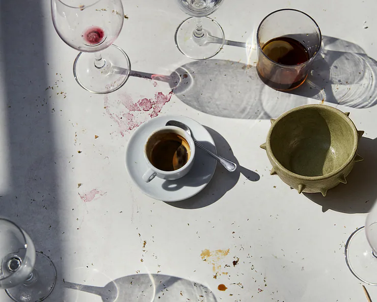 Tavolo imbandito con bicchieri e tazze usati, tra cui un espresso in tazzina bianca, calici di vino e ciotole verdi.