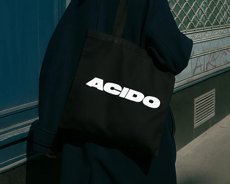 Persona con cappotto scuro porta una tote bag nera con la scritta bianca ACIDO in caratteri grandi e audaci.