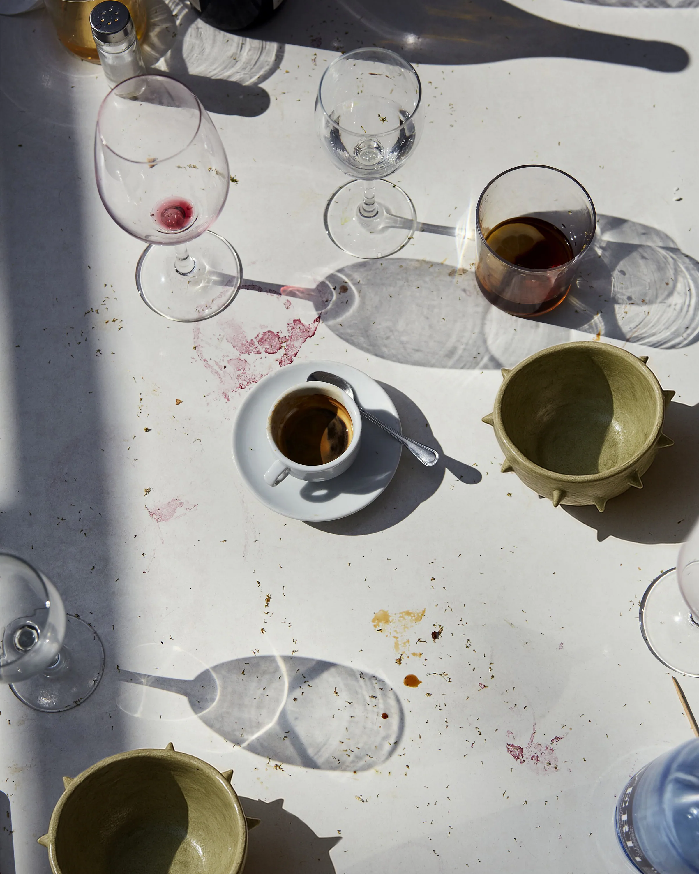 Tavolo con bicchieri sporchi di vino, tazzina di caffè e ciotole in ceramica verde, con macchie e ombre proiettate dalla luce del sole.