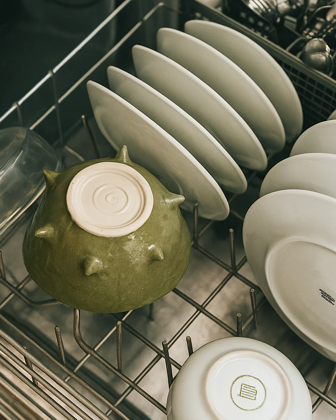 Lavastoviglie aperta con piatti bianchi ordinati e una ciotola verde in ceramica capovolta.