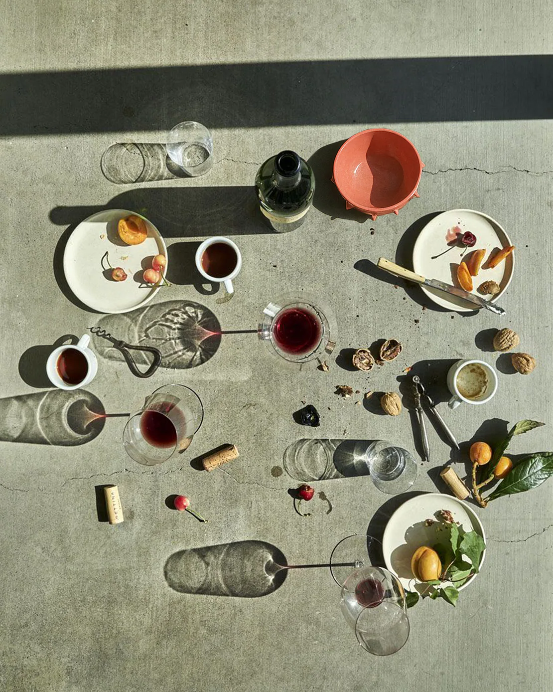 Tavolo in cemento con bicchieri di vino, tazze da caffè, piatti con frutta e noci, e una ciotola rossa in ceramica.