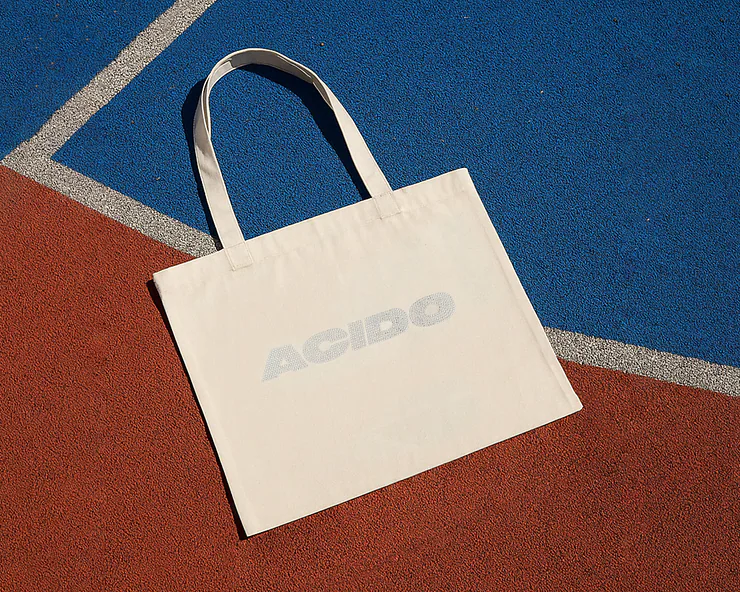 Shopper in tessuto bianco con scritta ACIDO appoggiata su un campo sportivo con fondo blu e rosso.