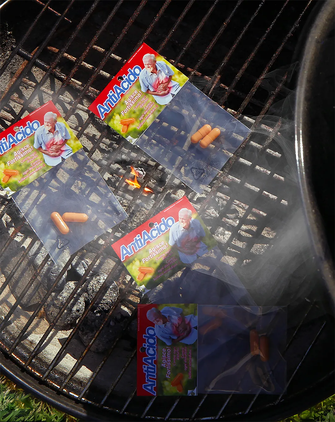 Confezioni di Antiacido Pills posizionate su una griglia da barbecue con fiamme e fumo, creando un'ambientazione ironica e surreale.