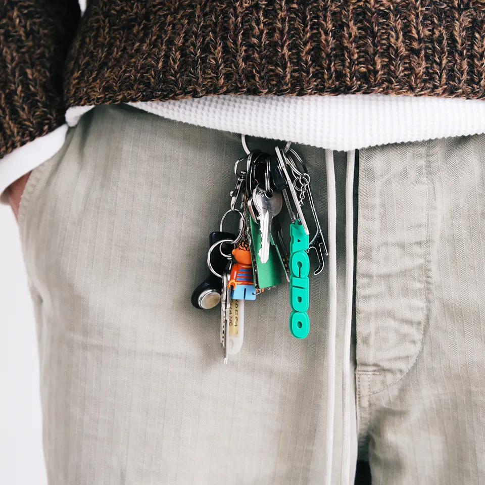 Portachiavi verde 'ACIDO' appeso a un mazzo di chiavi e fissato a un paio di pantaloni in jeans.