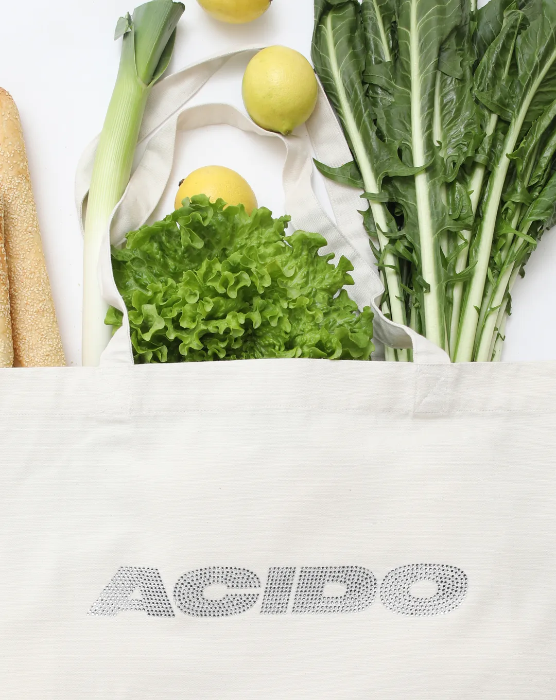 Dettaglio della Totebag White 'ACIDO' con logo in brillantini argentati, riempita con insalata, limoni, pane e verdure fresche.