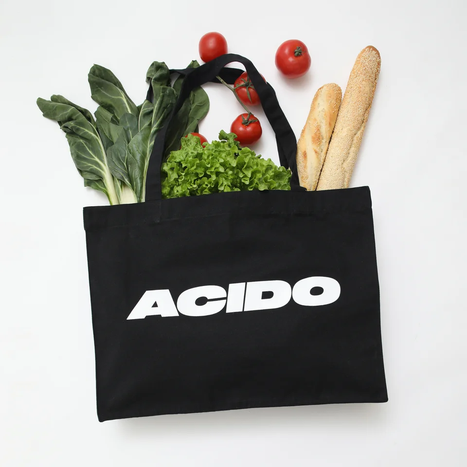 Tote bag nera con il logo 'ACIDO' riempita con verdure fresche come lattuga, coste e pomodori.