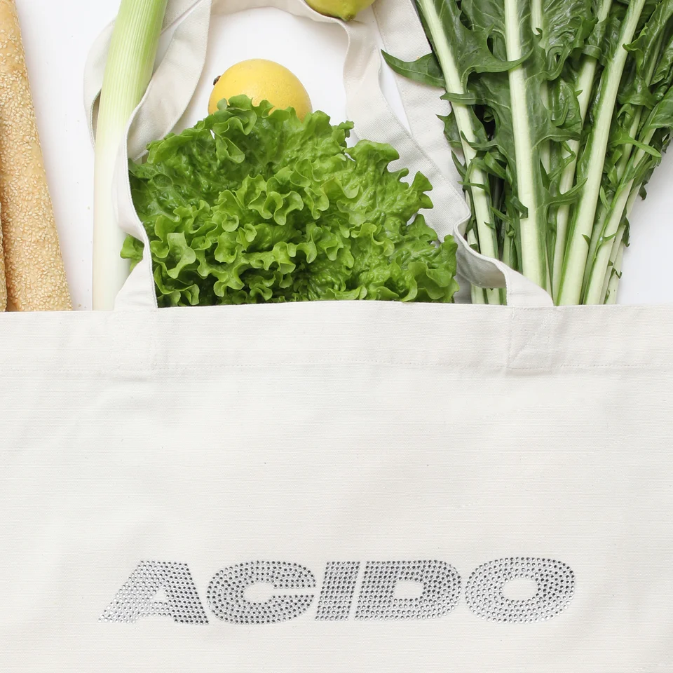 Dettaglio della Totebag White 'ACIDO' con logo in brillantini argentati, riempita con insalata, limoni, pane e verdure fresche.