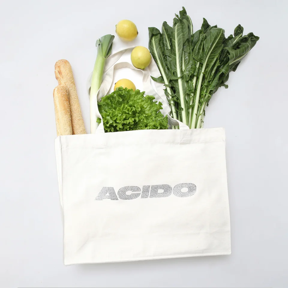 Totebag bianca 'ACIDO' riempita con pane, limoni, insalata e verdure fresche, ideale per lo shopping quotidiano.