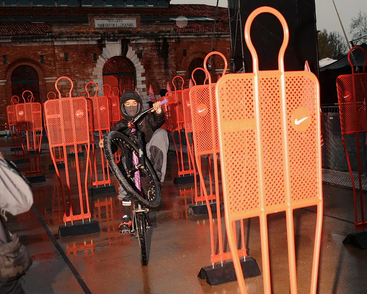 Ciclista che esegue un’impennata tra sagome da allenamento arancioni con logo Nike, in un cortile urbano con edificio storico sullo sfondo.