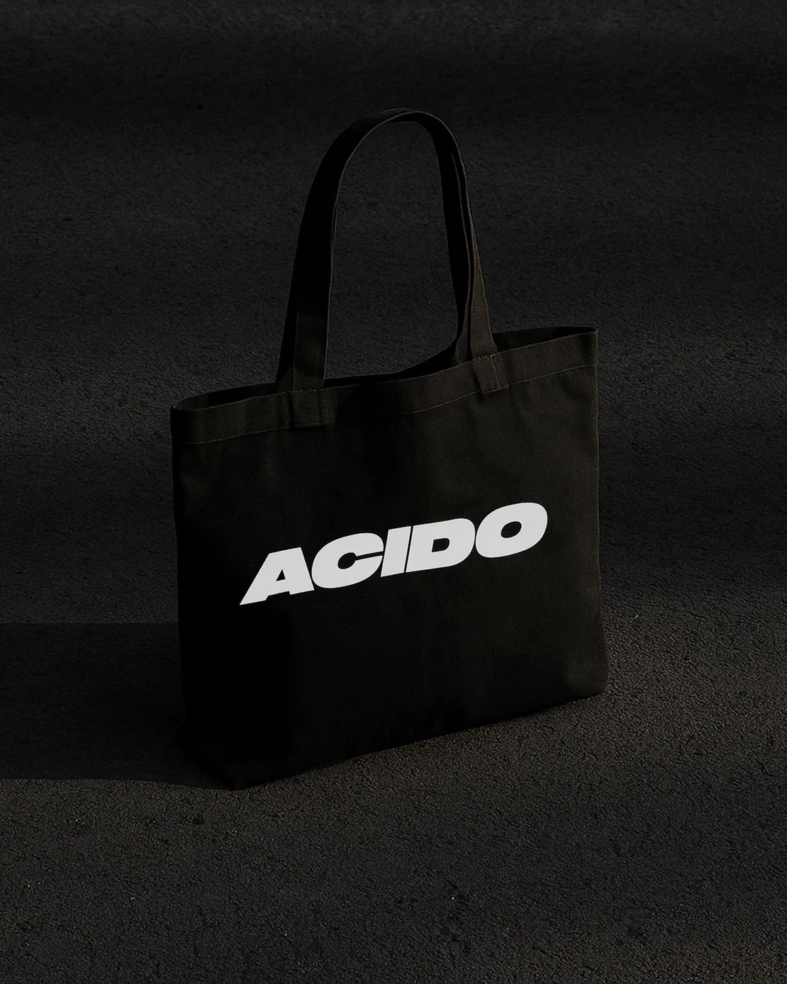 Totebag nera con scritta bianca ACIDO poggiata su asfalto scuro con luce e ombre nette.