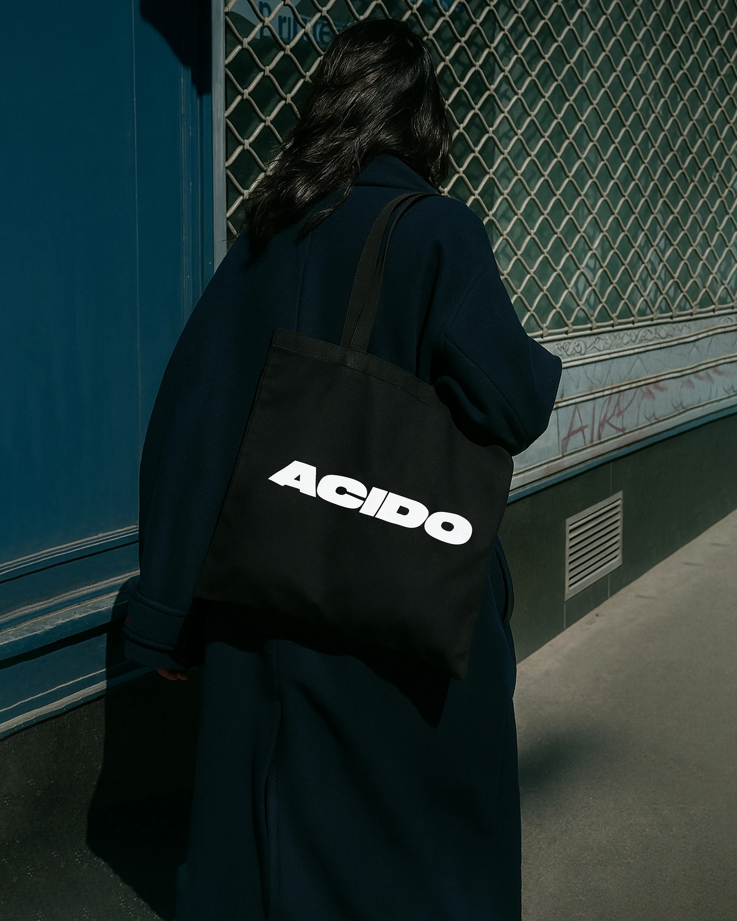 Totebag nera con scritta bianca ACIDO indossata da una persona con cappotto blu in città.