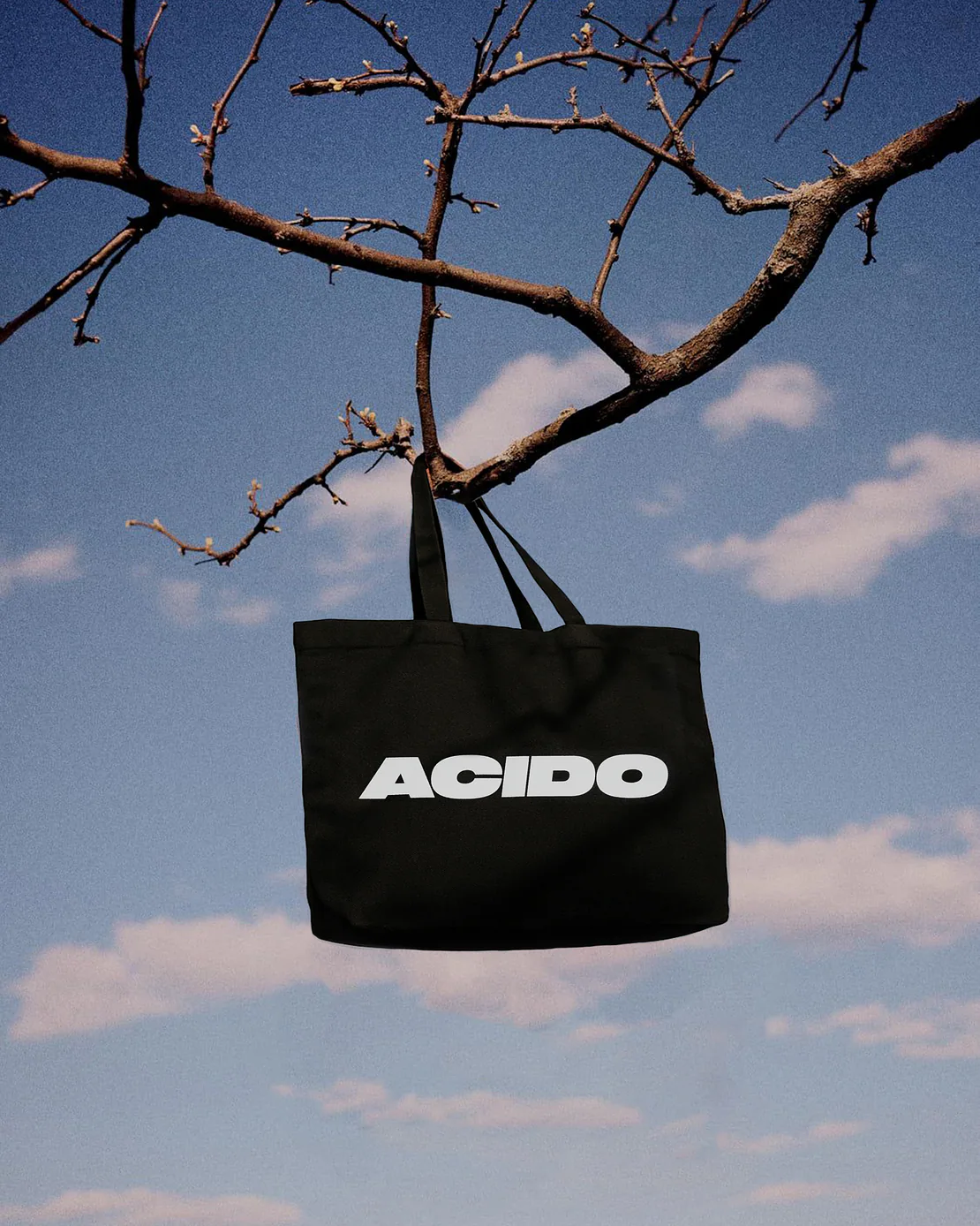 Totebag nera con scritta bianca ACIDO appesa a un ramo secco con cielo blu sullo sfondo.