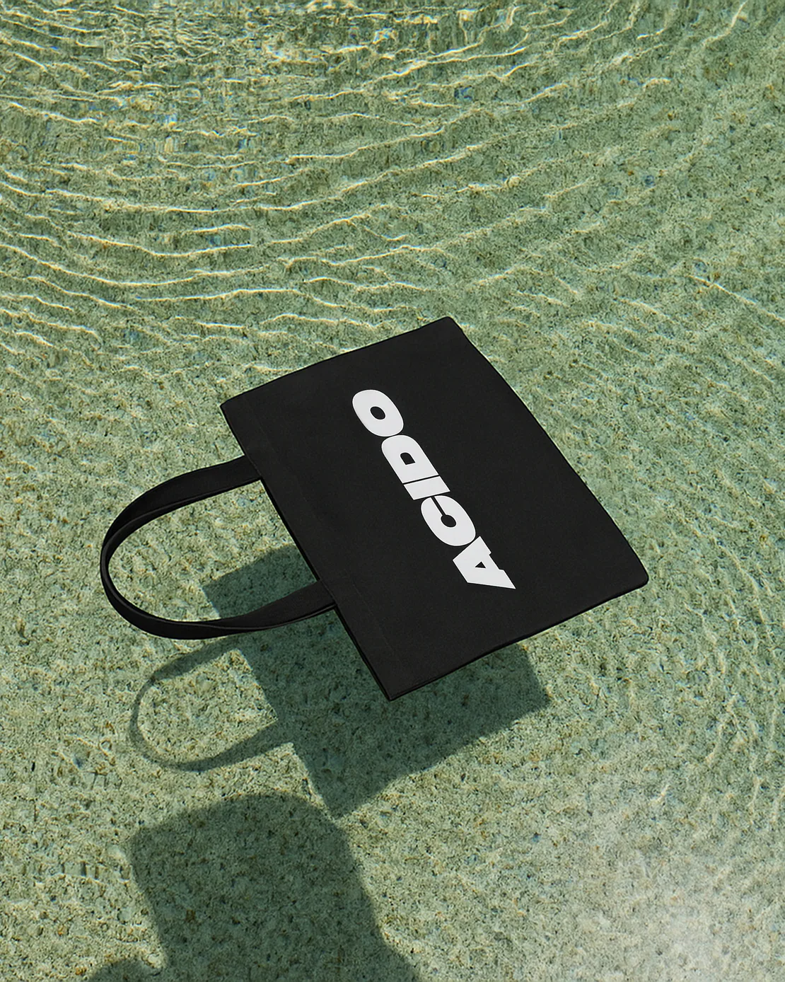 Totebag nera con scritta bianca ACIDO galleggiante sull’acqua limpida con riflessi di luce.