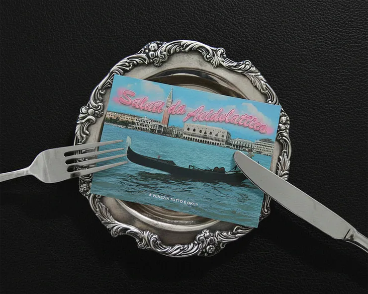 Cartolina con la scritta ‘Saluti da Acidolattico’ raffigurante Venezia, posata su un piatto d’argento con forchetta e coltello.