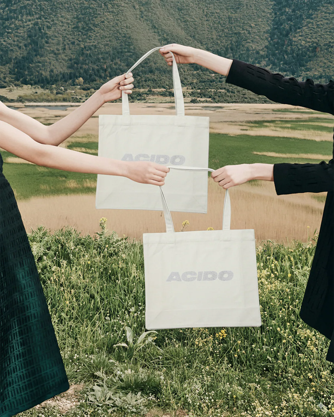 Due persone che tengono in mano due totebag bianche ACIDO davanti a un paesaggio naturale verde.