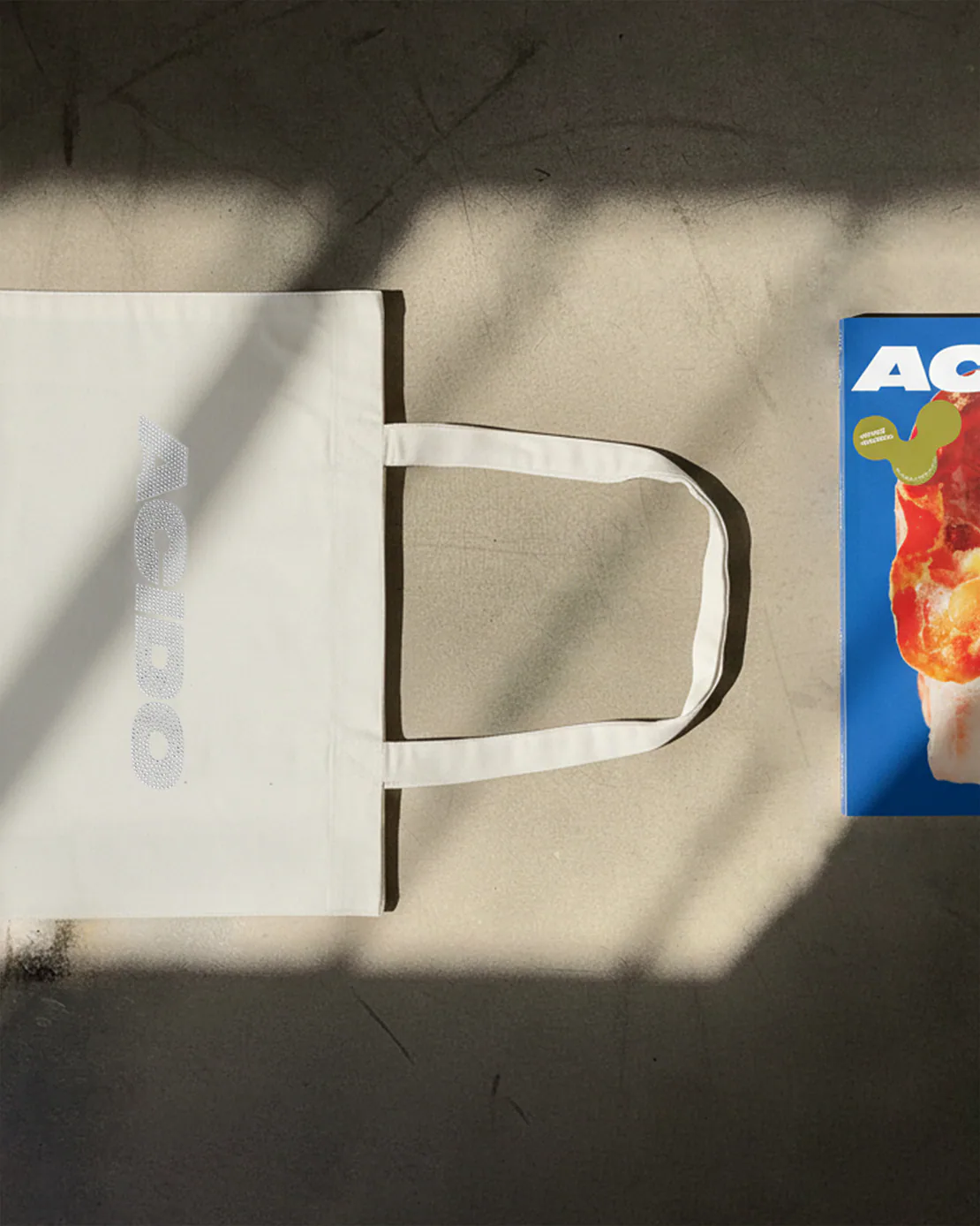 Totebag bianca ACIDO distesa a terra accanto a una rivista con copertina blu.