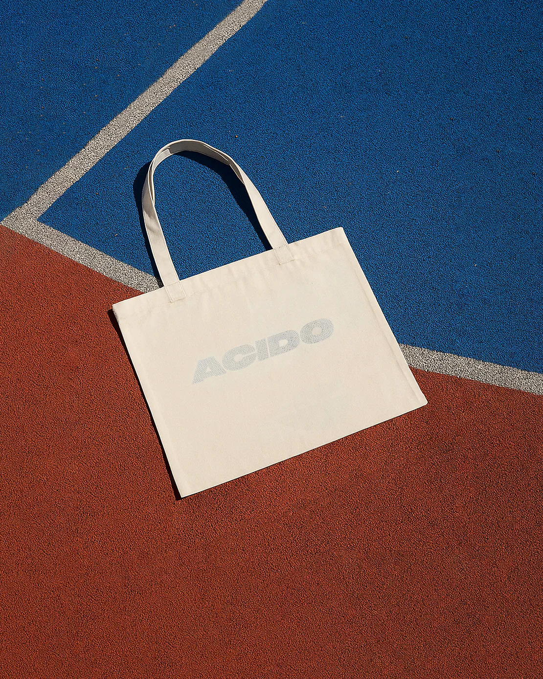 Totebag bianca ACIDO posata a terra su un campo sportivo con superfici rosse e blu.
